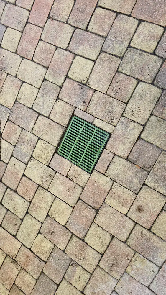A square green drain grate set flush into a herringbone brick paver surface