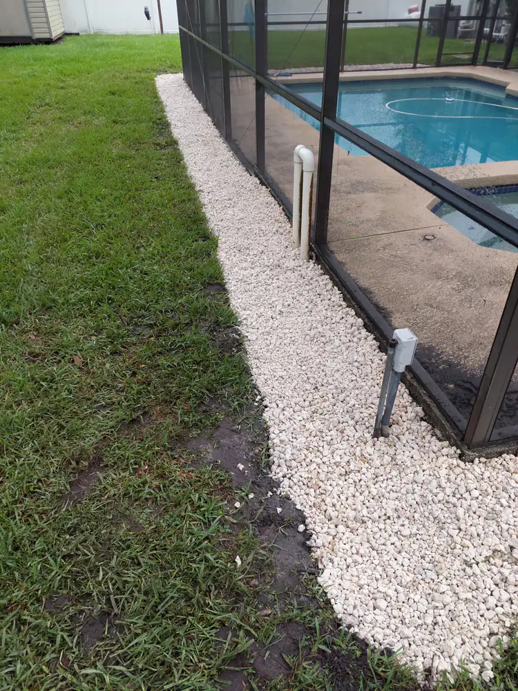 A white gravel drainage strip along a pool screen enclosure with a PVC riser