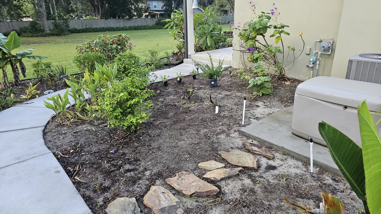 A bare dirt bed with flagstones staged and potted plants awaiting installation