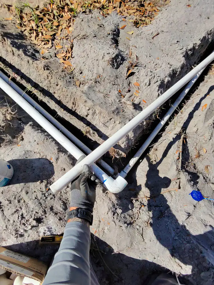 A gloved hand fitting a PVC elbow onto an irrigation pipe over an open trench