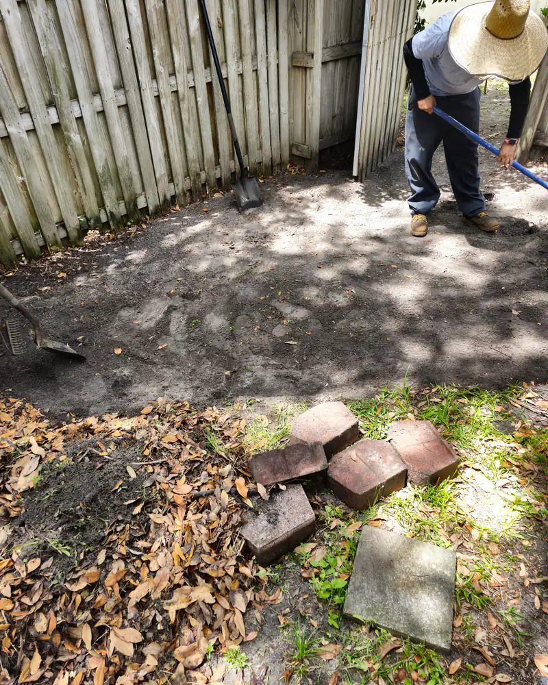 A Woods crew member in a straw hat grading and raking a Florida side yard by hand