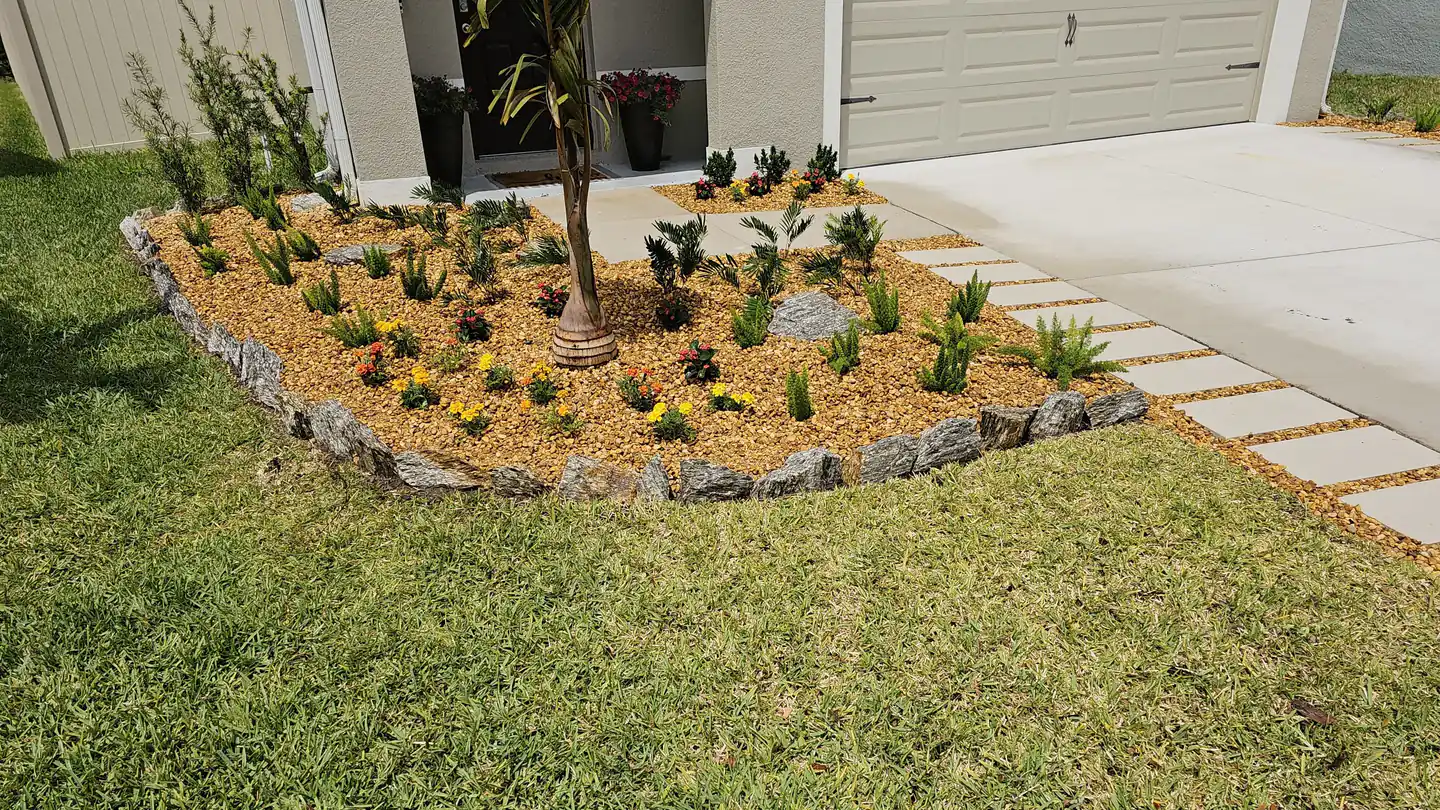 A front entry bed of gold river rock edged with stone, planted with a palm, dwarf conifers and marigolds, beside a stepping stone strip along the driveway