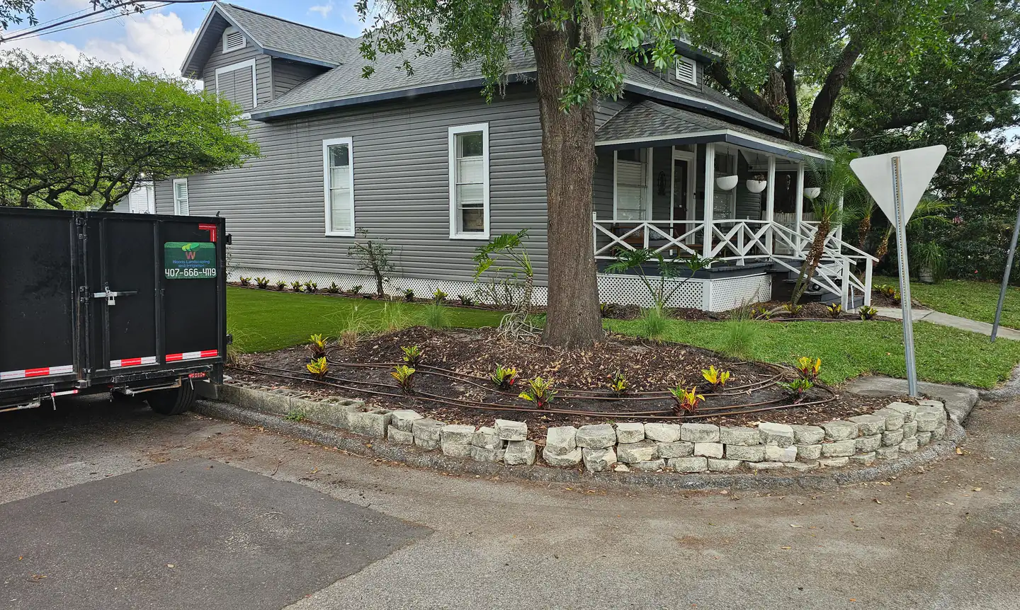 A Woods dump trailer parked at the kerb beside a finished corner-lot bed - a stone retaining wall, fresh mulch, new drip line and crotons, with a mown lawn and a covered porch behind