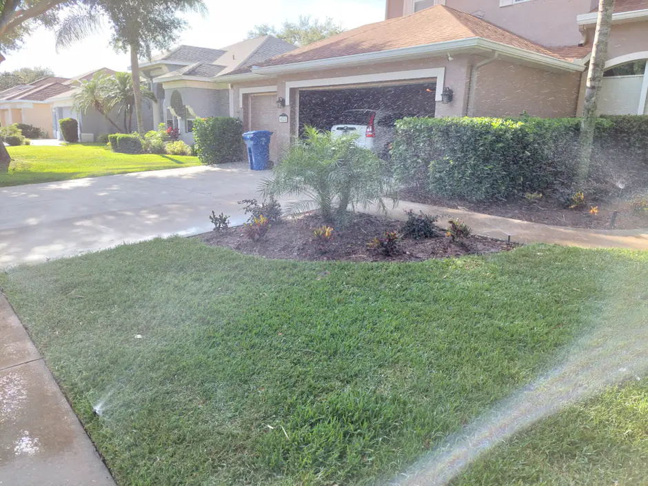 A Woods irrigation system running - sprinkler heads throwing arcs of water across a fresh green front lawn beside a new palm and croton bed